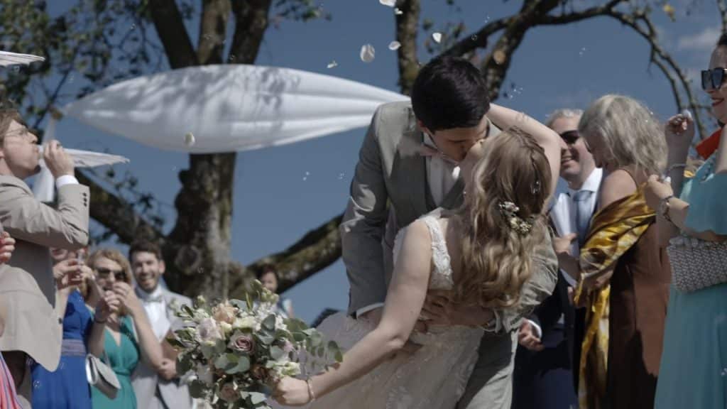 "Ein Brautpaar teilt einen innigen Kuss bei ihrer Hochzeit im Freien, umgeben von jubelnden Gästen, die Seifenblasen blasen und Blumenblätter werfen. Die Braut trägt ein weißes Spitzenkleid und hält einen Blumenstrauß, während der Bräutigam einen grauen Anzug trägt. Im Hintergrund sind weiße Dekorationen und ein blauer Himmel zu sehen."